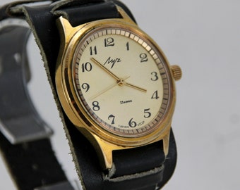 May include: A gold-tone wristwatch with a black leather band. The watch face is white with black numerals and a 'Mir' inscription. The watch face also has the text '23 камня' and 'Сделано в СССР'.