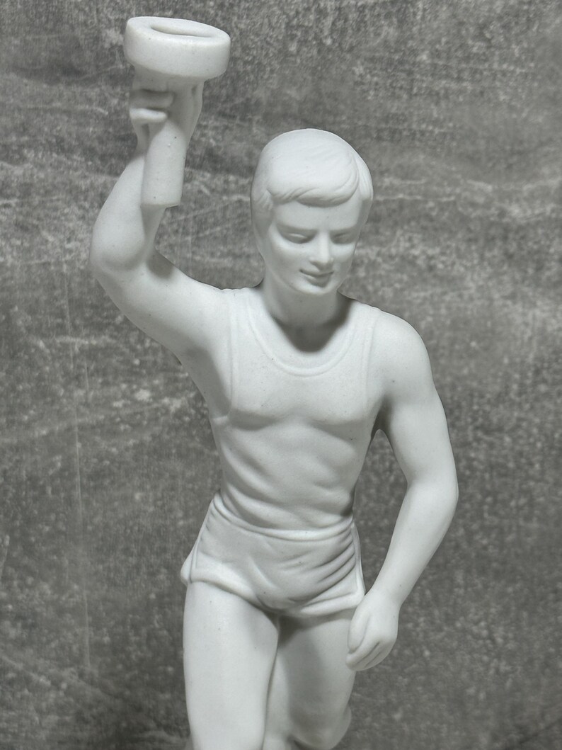 May include: White porcelain statue of a runner holding a torch. The runner is wearing a sleeveless top and shorts. The statue is highly detailed and shows the runner in mid-stride.  The piece is a collectible figurine, perfect for sports enthusiasts or those who appreciate fine craftsmanship.