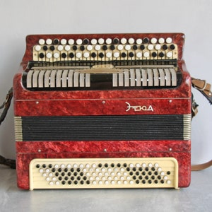 Puede incluir: Un acordeón rojo con teclas blancas y negras y un fuelle blanco y negro. El acordeón tiene una correa y la palabra "ELOZA" escrita en la parte delantera.