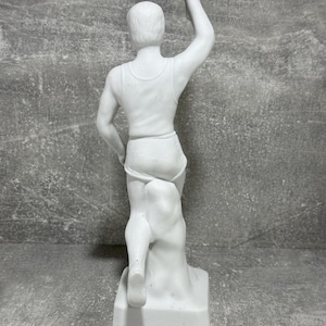 May include: White porcelain statue of a runner holding a torch. The statue is detailed, showing the runner's musculature and the texture of the fabric. The runner is depicted in a dynamic pose, suggesting movement and speed. The base of the statue is square and simple, providing a contrast to the more intricate details of the figure. The overall color is a uniform white, suggesting a clean and classic aesthetic. The statue is a collectible item, suitable for display in a home or office.