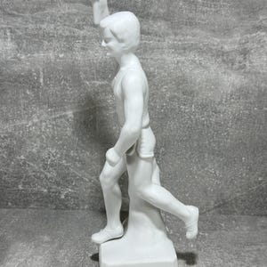 May include: White porcelain statue of a runner holding a torch. The statue is dynamic, capturing the runner in mid-stride. The figure is detailed, with visible musculature and facial features. The base is simple and rectangular, providing a stable platform for the piece. The overall style is classic and timeless, suitable for display in a home or office setting.  The statue is a collectible item, possibly representing athletic achievement or victory.