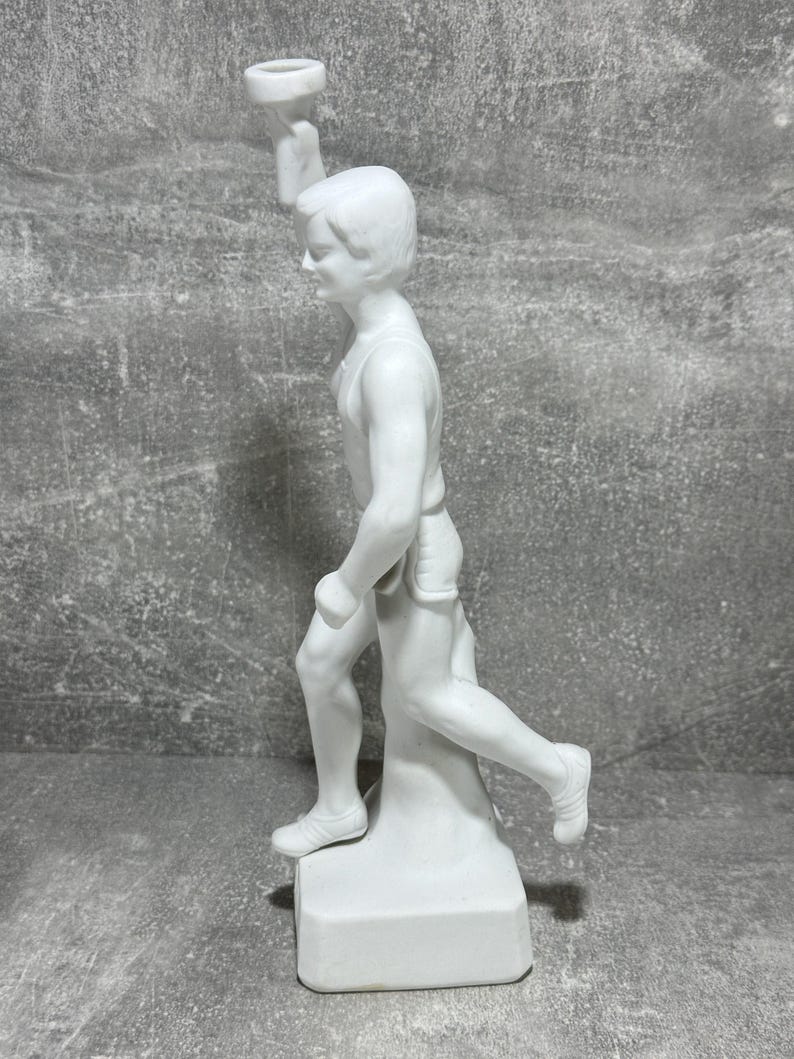 May include: White porcelain statue of a runner holding a torch. The statue is dynamic, capturing the runner in mid-stride. The figure is detailed, with visible musculature and facial features. The base is simple and rectangular, providing a stable platform for the piece. The overall style is classic and timeless, suitable for display in a home or office setting.  The statue is a collectible item, possibly representing athletic achievement or victory.