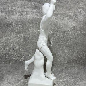 May include: White porcelain statue of a runner holding a torch. The runner is depicted mid-stride, with one leg raised higher than the other. The statue is mounted on a square base. The figure is detailed and shows the musculature of the runner. The statue is a collectible item and is suitable for display in a home or office.