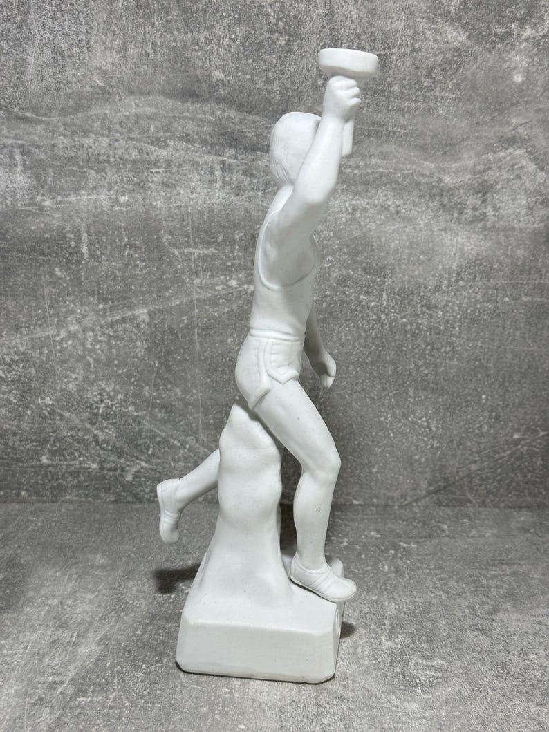 May include: White porcelain statue of a runner holding a torch. The runner is depicted mid-stride, with one leg raised higher than the other. The statue is mounted on a square base. The figure is detailed and shows the musculature of the runner. The statue is a collectible item and is suitable for display in a home or office.