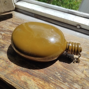 May include: A green metal canteen with a screw-on cap and a chain attached. The canteen is sitting on a wooden surface.