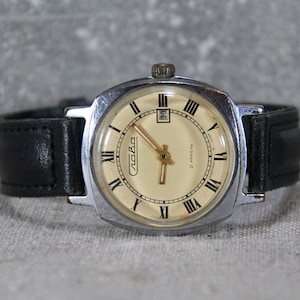 May include: A silver-toned wristwatch with a square face and black leather band. The watch face has Roman numeral hour markers and a date window at the 3 o'clock position. The watch is labeled "Слава" and "21 JEWELS".