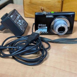 May include: A black Nikon Coolpix digital camera with a silver lens, a charger, and a USB cable. The camera has a Scene Auto Select feature and 4x zoom. The camera is on a wooden surface.
