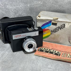 Puede incluir: Una cámara de película de 35 mm negra y plateada con una funda de cuero. La cámara tiene una inscripción roja que dice "SMENA 8M". La cámara está sobre una superficie gris con una caja y un manual de instrucciones.