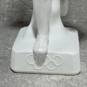 May include: White porcelain Olympic athlete figurine on a square base. The base features the Olympic rings emblem. The figurine depicts an athlete in mid-stride, wearing running shoes. The figure is detailed and shows musculature. The piece is a collectible item, possibly vintage.