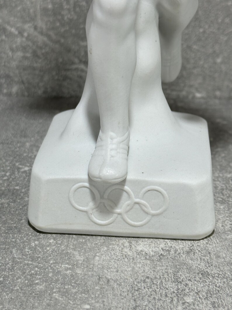 May include: White porcelain Olympic athlete figurine on a square base. The base features the Olympic rings emblem. The figurine depicts an athlete in mid-stride, wearing running shoes. The figure is detailed and shows musculature. The piece is a collectible item, possibly vintage.