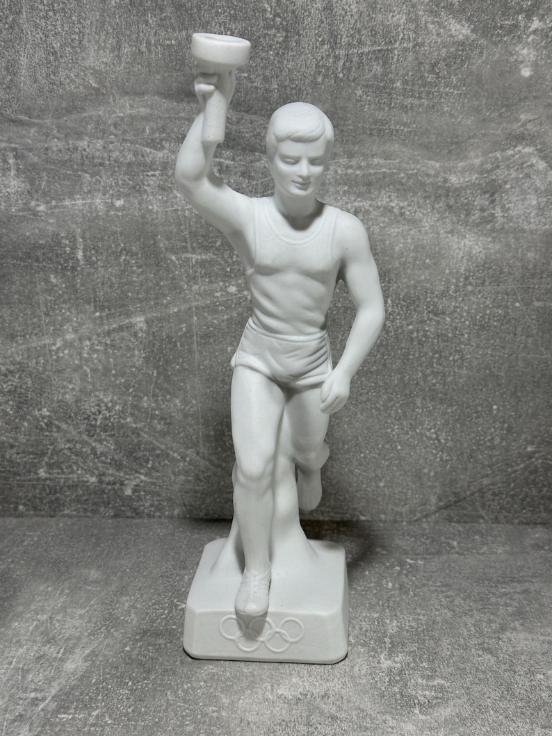 May include: White porcelain figurine of a runner holding a torch. The runner is depicted in a dynamic pose, mid-stride, with the torch held high. The figurine stands on a square base with the Olympic rings emblem.  The piece is monochromatic, showcasing a smooth, matte finish. The details are finely sculpted, capturing the movement and energy of the athlete. This collectible figurine is a celebration of athletic achievement and the Olympic spirit.