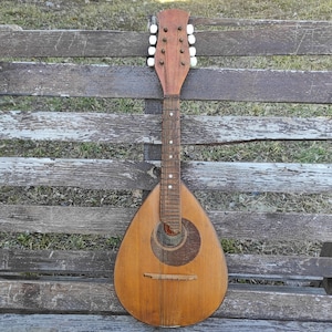 Puede incluir: Un mandolín de madera vintage con un acabado nacarado y clavijas de afinación blancas. El mandolín está apoyado en un banco de madera.