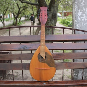 Puede incluir: Una mandolina de madera con un cuerpo marrón y un agujero de sonido marrón oscuro. La mandolina tiene un cuello marrón claro con una cabeza roja y clavijas de afinación negras.