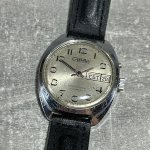 May include: A vintage silver-toned wristwatch with a black leather band. The watch face is silver with black numerals and hands. The watch face has the word "лава" and the day and date are visible.