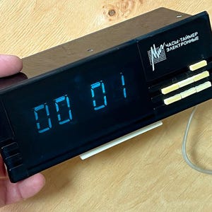 May include: A black digital clock with a blue display showing the time 00:01. The clock has a white label with the text "часы-таймер электронные" in Cyrillic script.