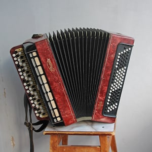 Könnte beinhalten: Ein rotes und schwarzes Akkordeon mit einem schwarzen Gurt und dem Wort "Sigma" auf der Seite. Das Akkordeon steht auf einem Holzhocker.