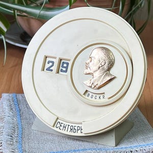 May include: Cream-coloured vintage calendar showing the date 29. A gold-coloured relief of a man's profile is centered on the dial, with the word "BOCKP" below. The month "Сентябрь" is printed at the bottom. The calendar sits on a grey and blue cloth.