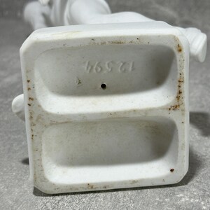 May include: White porcelain statue base with number 46571 imprinted. The base has two recessed areas.  The statue is of a classical style figure. The porcelain is matte white with some minor discoloration.