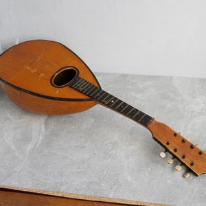 Könnte beinhalten: Eine vintage Holzmandoline mit braunem Korpus und einem schwarzen Streifen am Rand. Die Mandoline hat einen Holzhals und eine Reihe von Stimmwirbeln am Kopf.