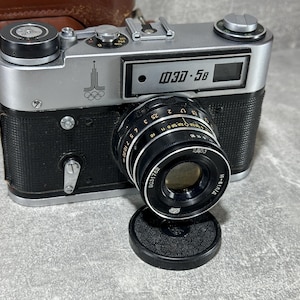 May include: A silver and black 35mm film camera with a black leather case. The camera has a lens with a black ring and a silver body. The camera has a black viewfinder and a silver shutter button. The camera has a black strap and a silver flash.
