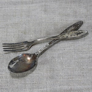 Peut inclure: Un ensemble de fourchette et cuillère en argent avec des motifs ornés. Les ustensiles sont posés sur une surface en lin gris.
