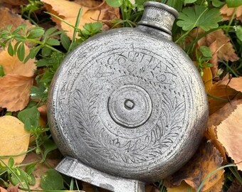 Könnte beinhalten: Eine silberfarbene, runde Flasche mit aufwendigen eingravierten Designs, darunter Text und florale Muster. Die Flasche hat einen kleinen, zylindrischen Hals und einen Sockel. Sie liegt auf einem Bett aus Herbstblättern und grünem Gras.