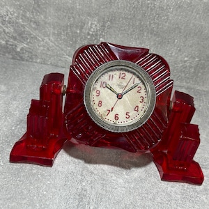 May include: A red plastic clock with a white face and black hands. The clock is on a red plastic stand shaped like a building.