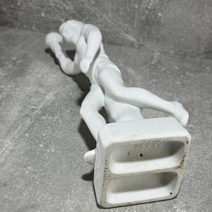 May include: White porcelain figurine of a male athlete in dynamic pose, possibly shot put or discus throw. The figure is sculpted with detail, capturing the movement and musculature. The base is square with recessed areas, and bears the number '76521'.