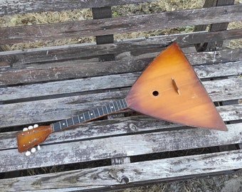 May include: A wooden triangular balalaika with a brown finish and a black fretboard. The instrument is lying on a weathered wooden surface.