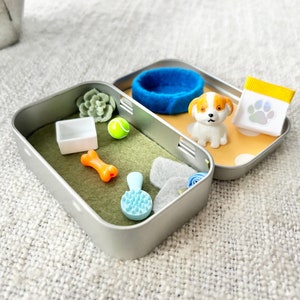May include: Miniature dog-themed accessories arranged in an open metal tin. Includes a small blue dog bed, a toy puppy, a bone, a brush, a tennis ball, and a bag of dog food. The tin is set on a textured surface.