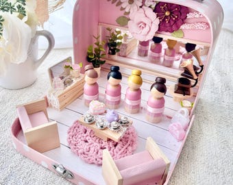 May include: A pink suitcase dollhouse interior featuring four wooden peg dolls in pink dresses, a miniature wooden table with tea accessories, two small pink chairs, a round pink crocheted rug, and floral wall decorations.