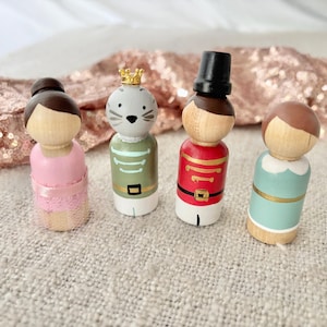 May include: Four wooden peg dolls. A ballerina in a pink dress, a cat with a crown, a soldier in a red uniform, and a person in a blue dress. The dolls are painted with details and stand on a textured surface.