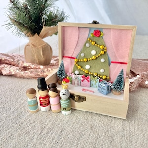 May include: A wooden Christmas-themed display box with a felt Christmas tree, miniature gifts, and decorative peg dolls. The box is open, revealing a pink backdrop and a small Christmas tree. The peg dolls include a nutcracker and a cat.