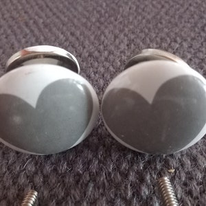 Op de afbeelding: Twee grijze keramische kastknoppen met een wit hartontwerp. De knoppen hebben een zilveren metalen basis.