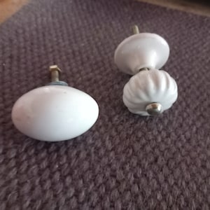 Pode incluir: Dois botões de armário de cerâmica branca. Um botão é redondo e o outro é de estilo mais tradicional com um design canelado.
