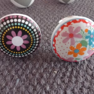 Könnte beinhalten: Zwei Keramik-Schubladengriffe mit Blumendesigns. Ein Griff hat einen schwarzen Hintergrund mit einer rosa Blume und einem Ring aus bunten Punkten. Der andere Griff hat einen weißen Hintergrund mit rosa, orangefarbenen und blauen Blumen.