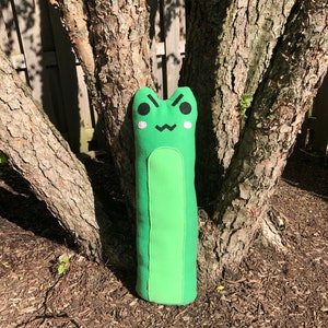 May include: A green plush frog pillow with a cute cartoon face. The frog has black eyes, a pink nose, and a black mouth. The pillow is standing upright against a tree trunk.