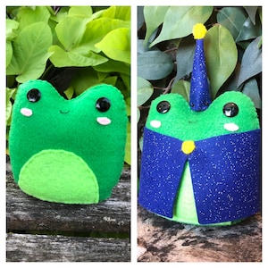 May include: Two green felt frog plushies. One frog is wearing a blue and yellow wizard hat and a blue cape with silver glitter.