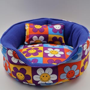 May include: A round pet bed featuring a vibrant checkerboard pattern of colourful flowers with smiley faces in shades of purple, orange, yellow, blue, and white. The interior is lined with solid purple fabric and includes a matching rectangular pillow.