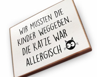 May include: White wooden sign with black text that reads "Wir mussten die Kinder weggeben. Die Katze war allergisch."  A black cat with white eyes is drawn on the sign.