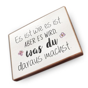 Peut inclure: Une pancarte en bois blanc avec un cadre brun et du texte noir qui dit "Es ist wie es ist. ABER ES WIRD, was du daraus machst." Il y a un papillon rose et une fleur rose sur la pancarte.