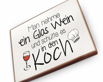 May include: White wooden sign with brown trim, featuring a black and white illustration of a wine glass and a chef's hat. The text reads "Man nehme ein Glas Wein und schütte es in den Koch".