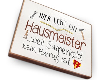 May include: White wooden sign with brown trim, featuring the text "Hier lebt ein Hausmeister...weil Superheld kein Beruf ist." with illustrations of tools, a paintbrush, a measuring tape, and a superhero logo.