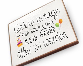 May include: White wooden sign with black text that reads "Geburtstage sind noch lange kein Grund älter zu werden". The sign is decorated with a birthday cake and balloons.
