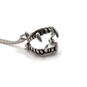 Puede incluir: Un collar con colgante plateado que presenta un diseño detallado de mandíbula abierta con colmillos y dientes afilados. El colgante cuelga de una cadena fina, creando un accesorio gótico o temático de Halloween.