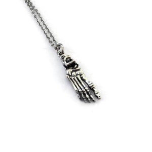 May include: A silver pendant necklace with a detailed skeletal foot charm. The charm hangs from a delicate chain, creating a gothic aesthetic. The necklace is displayed against a white background.