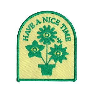 May include: Green embroidered patch with a yellow background. The patch features three stylized flowers in a pot with the text "HAVE A NICE TIME" around the flowers.