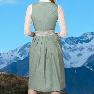 May include: A green dirndl dress with white polka dots, a white blouse, and lace sleeves. The dress has a light gray sash and is set against a backdrop of mountains and a blue sky. The traditional dress is a classic style.