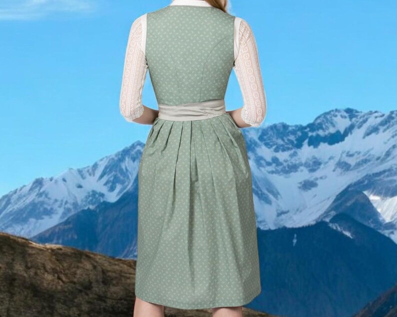 May include: A green dirndl dress with white polka dots, a white blouse, and lace sleeves. The dress has a light gray sash and is set against a backdrop of mountains and a blue sky. The traditional dress is a classic style.