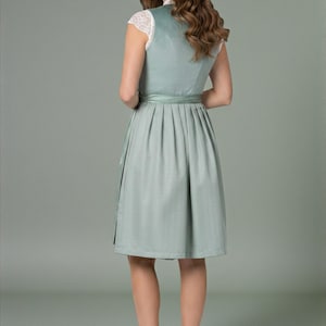 K&ouml;nnte beinhalten: Ein salbeigr&uuml;nes Dirndl mit &auml;rmellosem Oberteil, hochgeschlossenem Ausschnitt und einem Faltenrock. Das Kleid wird mit einer wei&szlig;en Spitzenbluse mit kurzen &Auml;rmeln, einem passenden Satin-Taillenband und beigen Pumps kombiniert.